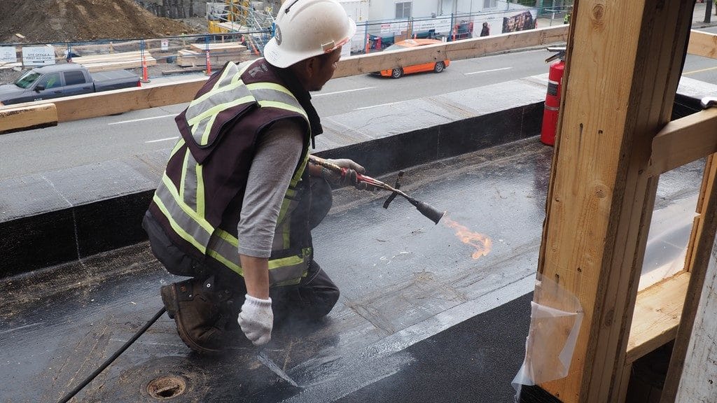 A construction worker in a hard hat and safety vest uses a torch to apply roofing material on a commercial job site. The urban setting is bustling with safety cones and equipment visible in the background.