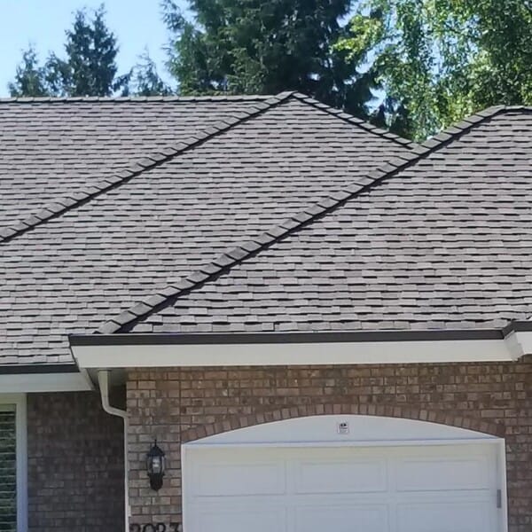 A brick house with a large garage featuring a white door and arched design. The roof, recently undergoing cedar roof replacement, boasts brown shingles. Two outdoor lights are mounted on either side of the garage. Trees and a clear sky are visible in the background.