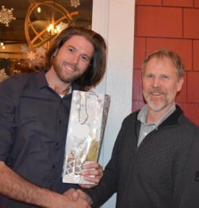 Two men are smiling and shaking hands indoors, celebrating client satisfaction. The man on the left, with long hair, holds a decorative gift bag. The man on the right sports a zip-up sweater. Holiday decorations in the background hint at the festive season atmosphere.