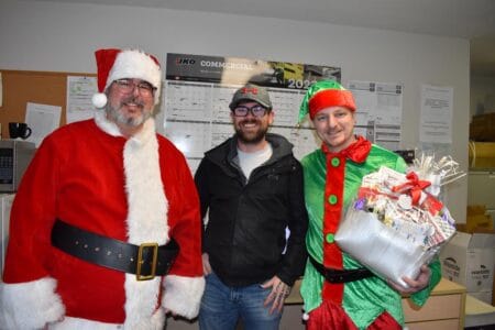 Two people dressed as Santa and an elf, with a gift basket in hand, stand beside a man in casual wear. The festive scene unfolds in an office where calendars and papers adorn the walls—a heartwarming celebration of client satisfaction during the joyous festive season.