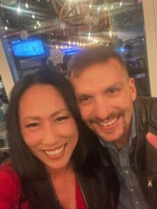 A woman and a man smile for a selfie in a cozy indoor setting, their warmth echoing the festive season. In the background, a TV displays a sports game, and decorative snowflakes adorn the window. Soft lighting enhances this cheerful moment of client satisfaction.