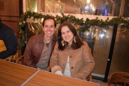 A man and woman, embodying client satisfaction, sit together at a wooden table, smiling at the camera. Theyre donned in brown jackets with a festive garland behind them, perfectly capturing the holiday seasons warmth.