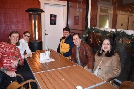 Five people are seated at an outdoor table with wooden surfaces, dressed warmly in jackets and sweaters. A tall heater is nearby, and festive garland decorates the space. Amid the festive season ambiance, a red wall and door frame their gathering, evoking warmth and charm.