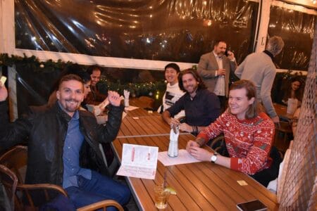 A group of people sits at a long wooden table in a lively festive season setting. Some are holding drinks, with one in a red sweater adorned with a Christmas pattern. The atmosphere exudes joy and relaxation.