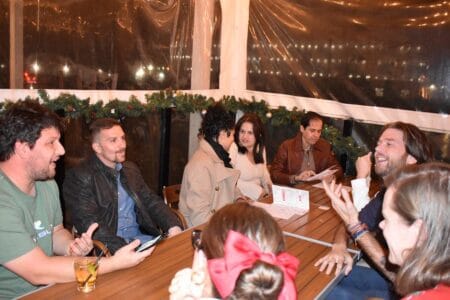A group of people sits around a wooden table, surrounded by garlands in a festive season atmosphere. They chat, laughing—one with a mobile phone, another enjoying a drink. Transparent plastic walls reveal blurred outdoor lights, fostering client satisfaction in every detail of their evening.