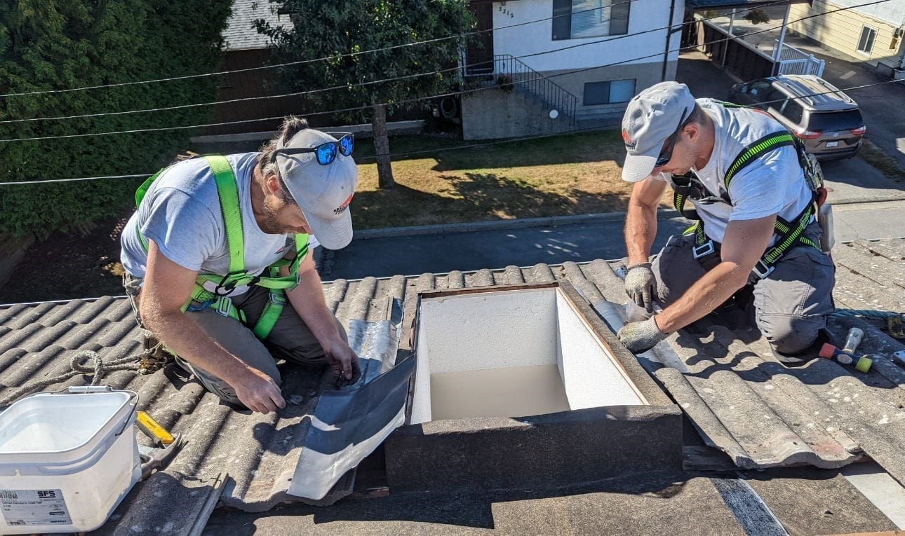 Two workers in safety harnesses and gray hats, from a roofing company in Vancouver, install or repair a skylight on a tiled roof. They handle tools and materials under bright daylight with houses, trees, and a parked car visible below.