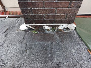 A close-up view of a brick chimney base on a shingled roof, showing common roofing issues such as deteriorating and cracked sealant, visible debris, and signs of water damage around the flashing—often noted by Vancouver home inspectors.
