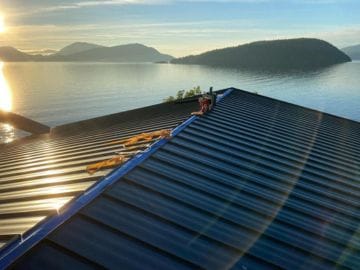 A rooftop with tools and gloves sits above a calm lake at sunset, ready for maintenance on metal roofs, with sunlight reflecting off the water and distant forested hills in the background.