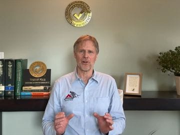 A man with light hair and a beard, wearing a light blue shirt, speaks while gesturing with his hands. He is seated in front of a shelf with books, a plant, and a plaque, discussing Absolute Roof Solutions, a roofing company in Greater Vancouver.
