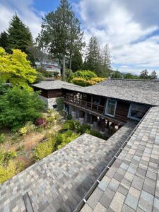 A view from a rooftop, featured in our residential roofing gallery, shows a house with grey shingles surrounded by lush green trees and shrubs under a partly cloudy sky. A garden area is visible next to the house.