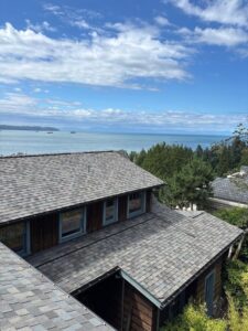 A wooden house with a gray shingle roof overlooks a scenic view of the ocean, green trees, and a partly cloudy blue sky. Ships are visible in the distant water, making it a perfect feature for any residential roofing gallery.