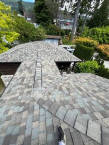 View from a rooftop with gray shingles, looking down at intersecting roof sections. A person’s shoe is visible at the bottom of the image. Trees and houses feature in the background of this residential roofing gallery scene.