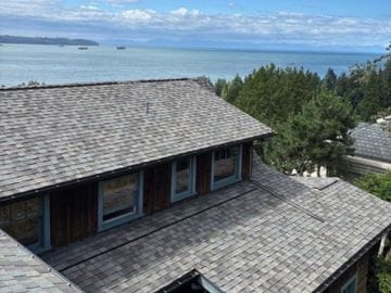 A West Vancouver home with a new CertainTeed Landmark shingle roof sits in the foreground, overlooking the ocean, trees, ships in the water, and a partly cloudy sky in the background.