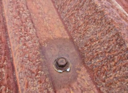 Close-up of a rusted corrugated metal sheet with a rusty bolt fastened in the center, illustrating the need for metal roof conversion in Richmond. The surface displays varying textures and shades of reddish-brown rust.