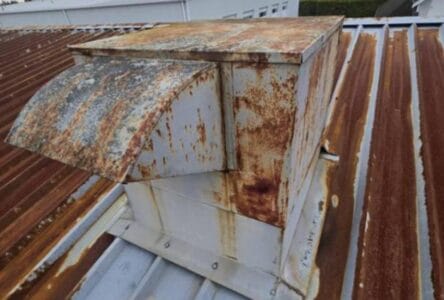 A rusted metal rooftop vent with significant corrosion sits on a corrugated metal roof in Richmond, also showing rust and weathering—an ideal candidate for a torch-on metal roof or upcoming metal roof conversion. Nearby buildings are partially visible.