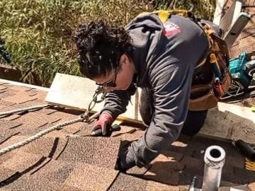 A person wearing a tool belt and safety gear is working on a shingled roof, using their hands to adjust or install shingles—a dedicated approach to roof repair with tools and ropes visible, ensuring quality roof solutions.