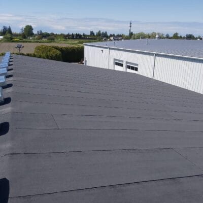 Flat black Torch-On Metal Roof atop a large industrial building with metal vents, adjacent to another structure in Richmond, surrounded by trees and open fields beneath a clear blue sky.