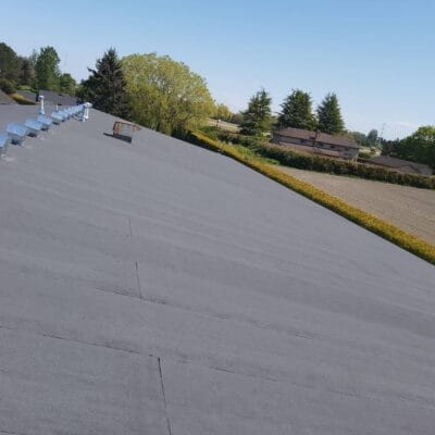 A wide, flat gray Torch-On Metal Roof with several vents is shown under a clear blue sky in Richmond, surrounded by trees, fields, and some houses in the background—ideal for a future Roof Conversion.