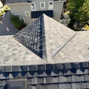 View from the top of a shingled roof looking down, showing intersecting roof peaks after a recent roof replacement, a vent, and a neighboring gray house with white trim and several trees in the background.