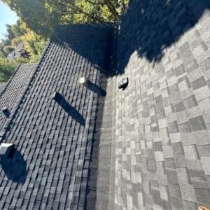 A close-up view of a dark gray asphalt shingle roof by Absolute Roof Solutions with a central valley, vent pipes, and vents, surrounded by trees casting shadows—ideal for housing cooperative roof replacement needs.