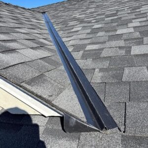 A close-up view of a house roof with dark gray shingles by Absolute Roof Solutions, featuring a metal flashing detail running along a valley where two roof slopes meet, under a clear blue sky.