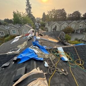 Workers from Absolute Roof Solutions repair a residential roof under a hazy sky with the sun visible overhead. Tools, tarps, and equipment are spread across the shingles of this housing cooperative, with townhouses and trees in the background.