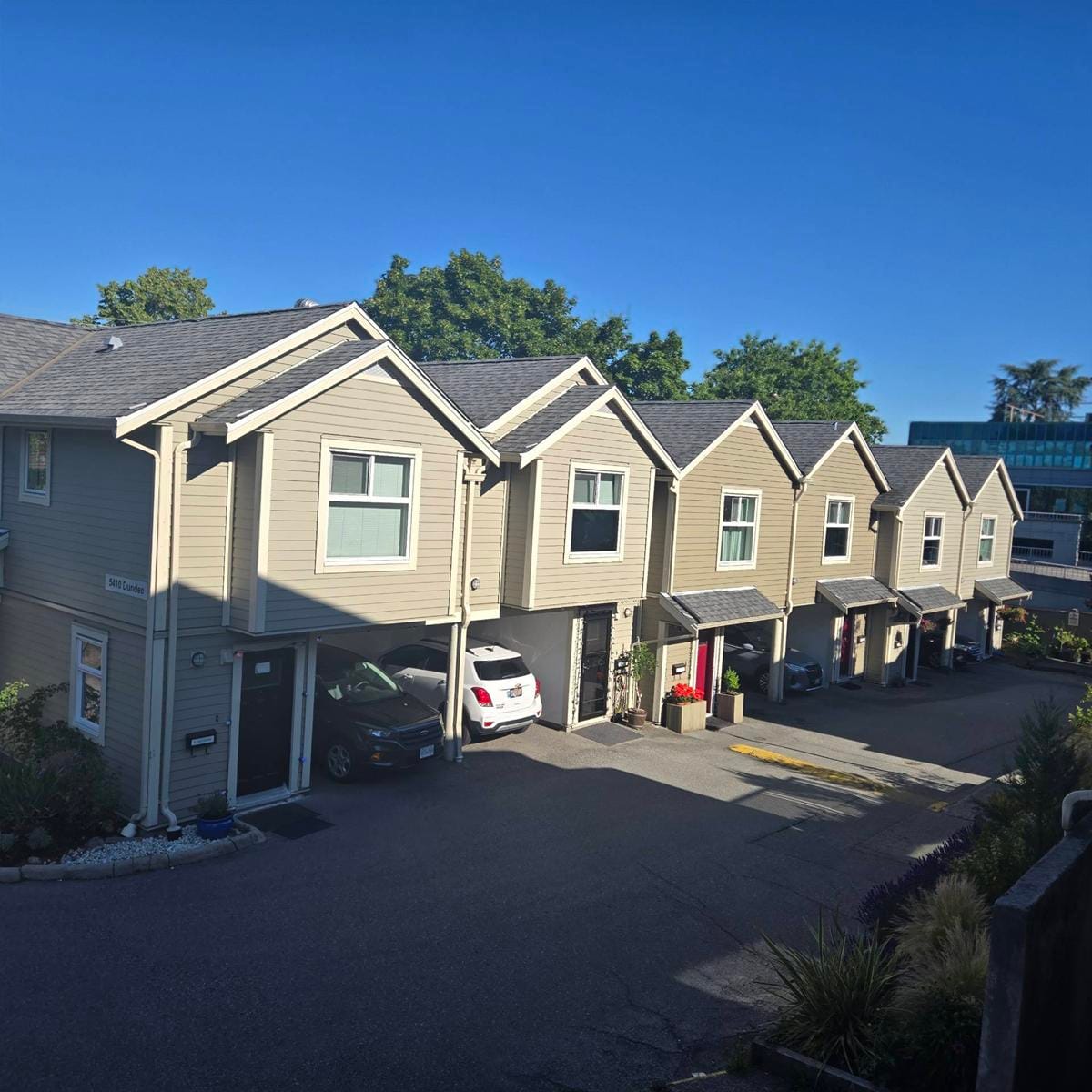 A row of modern, beige townhouses managed by a housing cooperative, with garages, parked cars, and greenery line a quiet residential street under a clear blue sky.