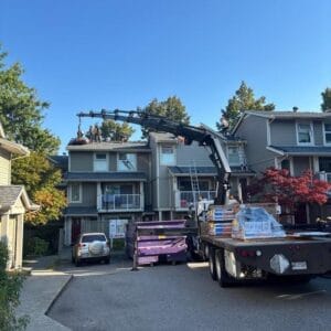 A truck with a mechanical crane from Absolute Roof Solutions lifts materials onto the roof of a townhouse, where workers handle a roof replacement. Several homes and trees are visible under a clear blue sky.
