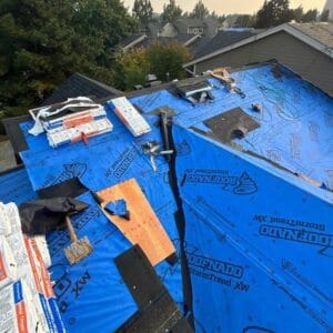 A house roof under construction, covered with blue underlayment sheets and tools scattered around. Roofing materials and equipment from Absolute Roof Solutions are placed on the roof, with trees and other houses visible in the background.